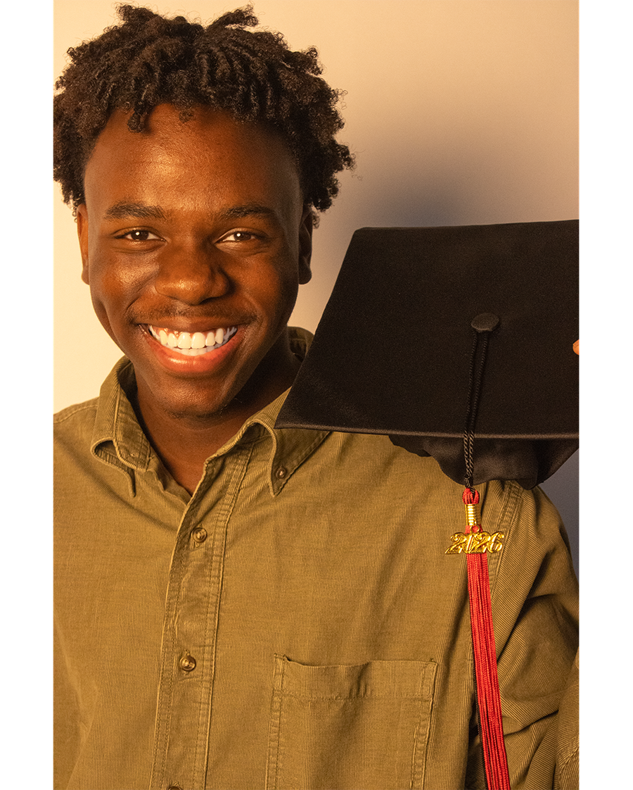 Close-up of the graduate laughing in an olive corduroy shirt, holding a cap with its red 2026 tassel beside their face, on a warm neutral backdrop