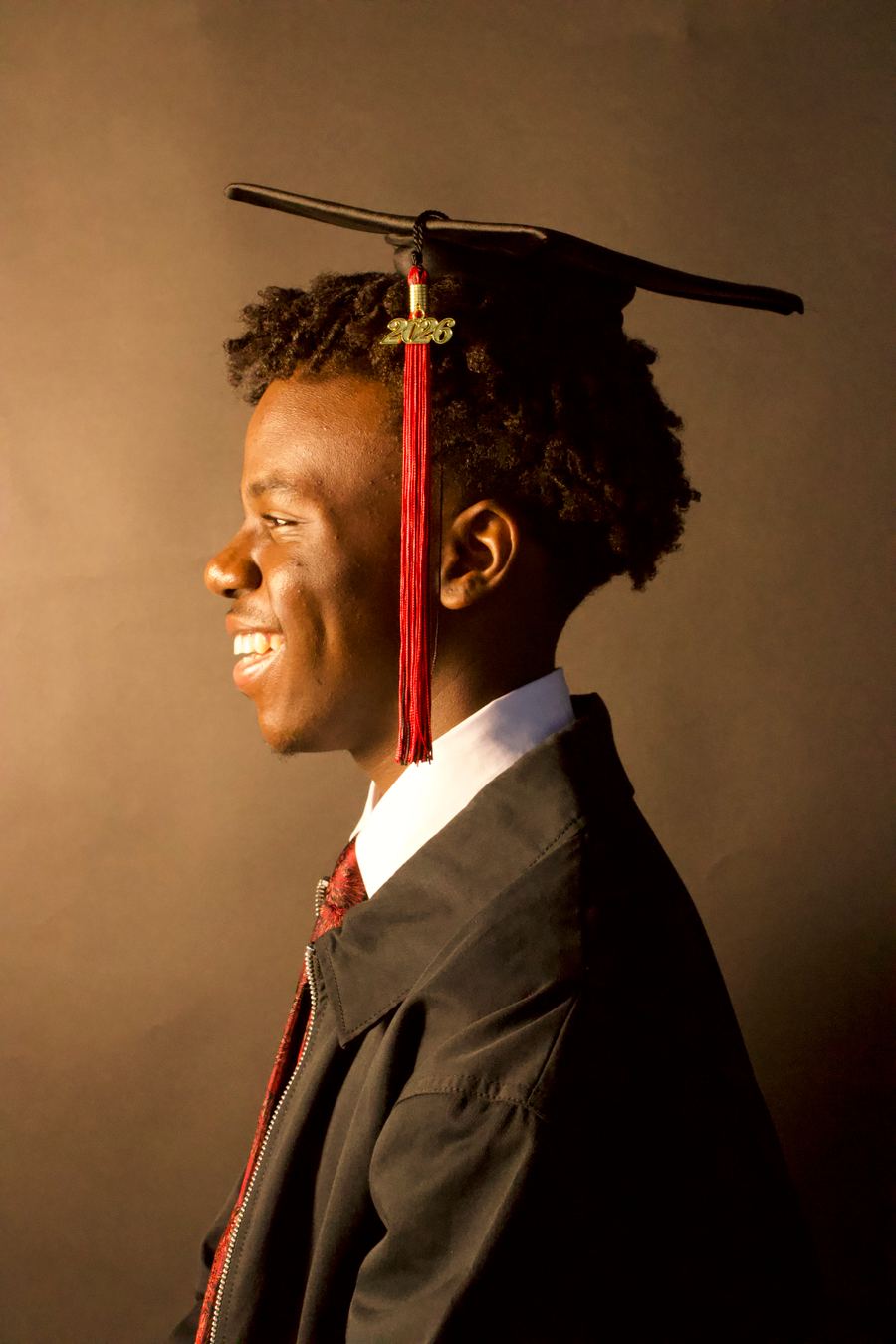 Side profile in cap, white collar and red tie against a dark brown backdrop, lit warmly from the front