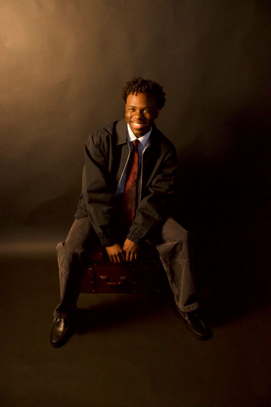 Full-length portrait of the graduate seated on a vintage suitcase in a dark jacket, red tie, jeans and loafers, smiling under a soft spotlight