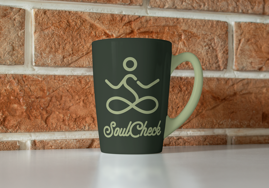 Dark green mug with a sage handle, printed with the SoulCheck meditating-figure mark and script wordmark, against a brick wall